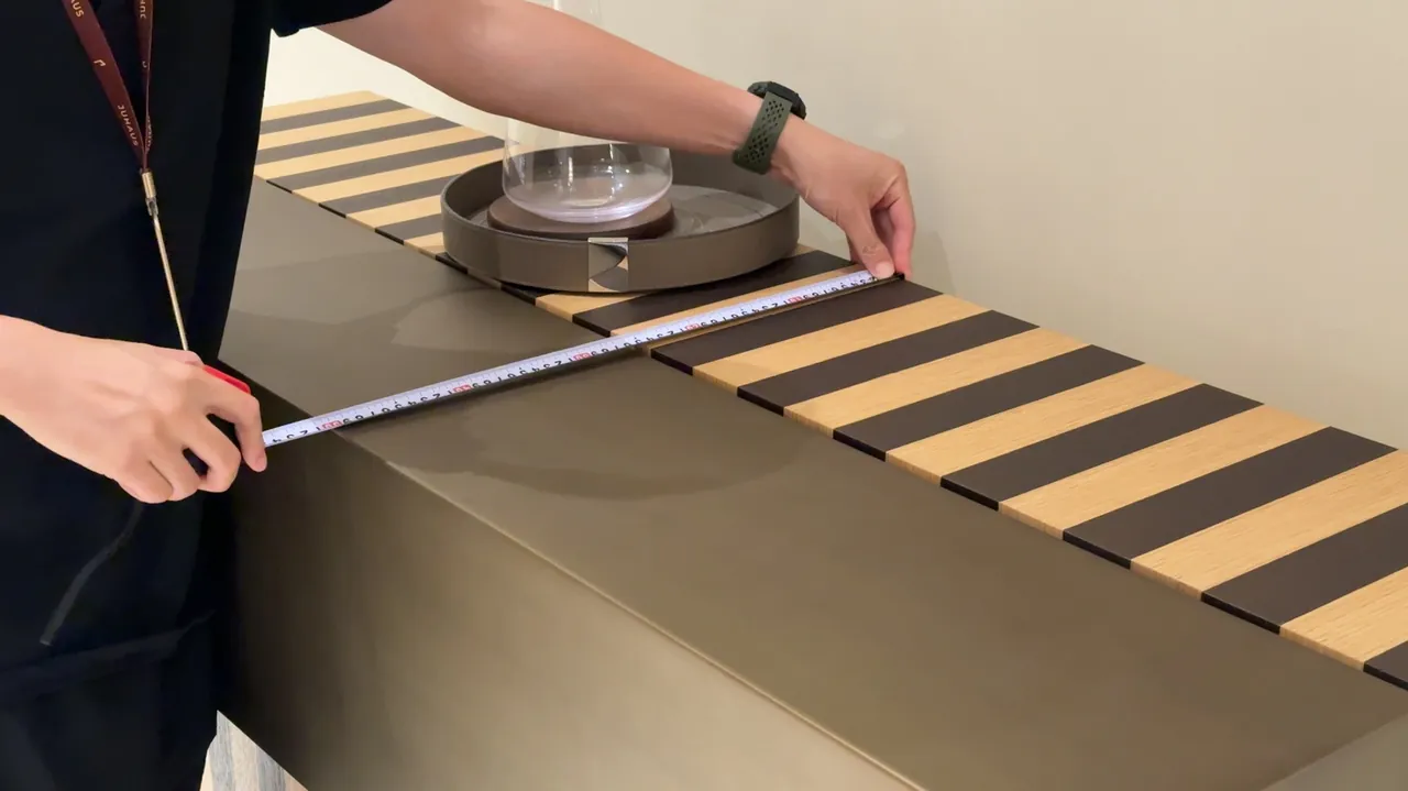 Hands stretch a tape measure across a furniture surface.