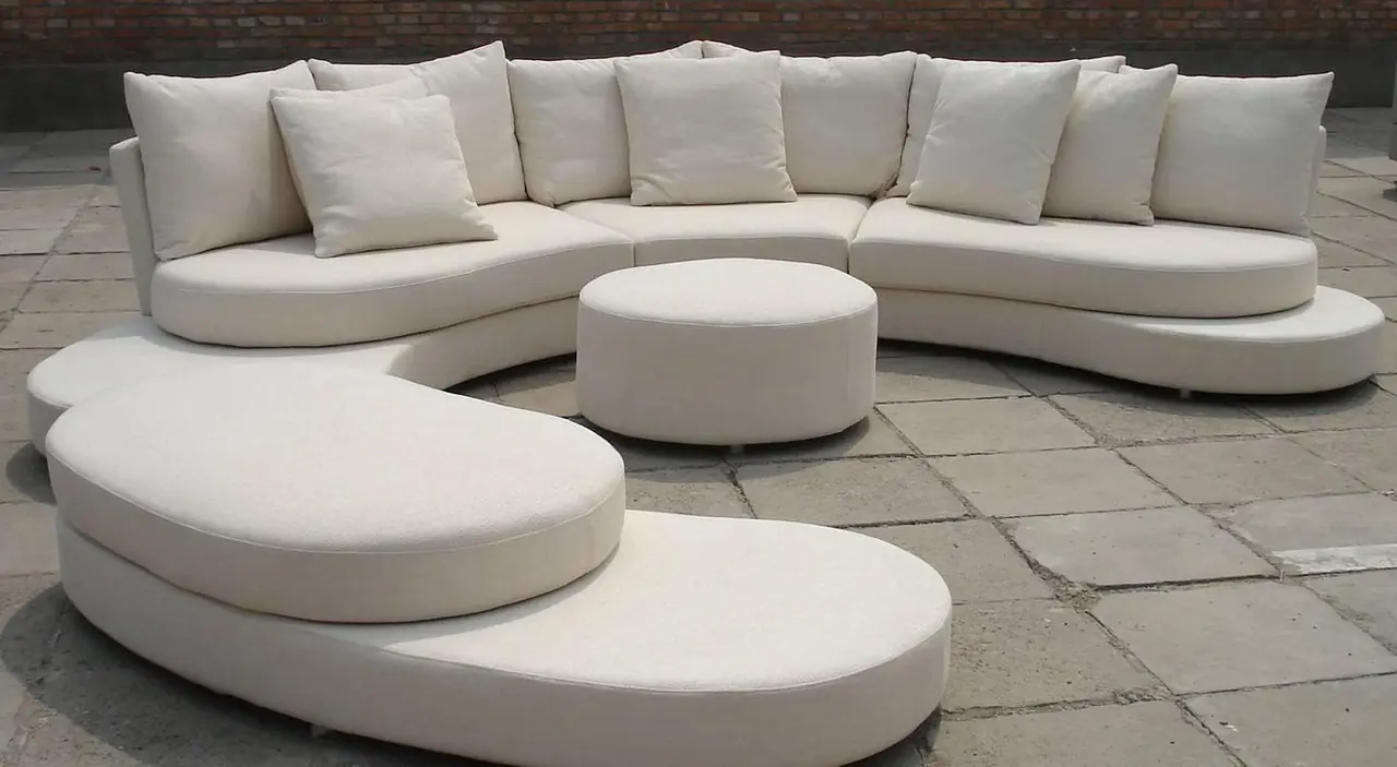 A low cream modular sofa in a curved arrangement with round ottoman on a paved setting.